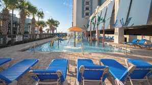 A pool area with blue lounge chairs, a water feature, palm trees, and a building with balconies.