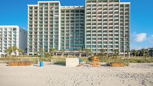 A beachfront hotel with multiple high-rise buildings, a sandy beach in the foreground, and palm trees.