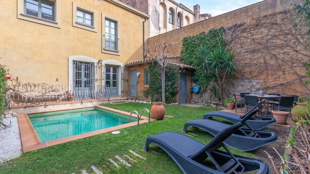 Piscina com espreguiçadeiras, mesa no alpendre e jardim com plantas e um muro de pedra.