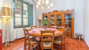 Uma sala de jantar com mesa redonda, cadeiras, um candelabro, um aparador com portas de vidro e uma janela com cortinas.
