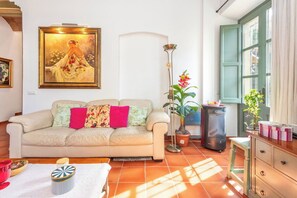 A living room with a sofa, a painting, a floor lamp, a potted plant, and a window with curtains.