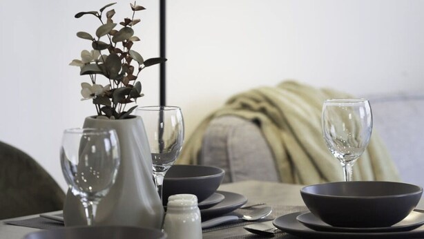 A dining table set with black bowls, plates, and wine glasses, accompanied by a vase with dried flowers.