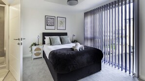 A modern bedroom with a large bed, a bedside table, a nightstand, and a window with vertical blinds.