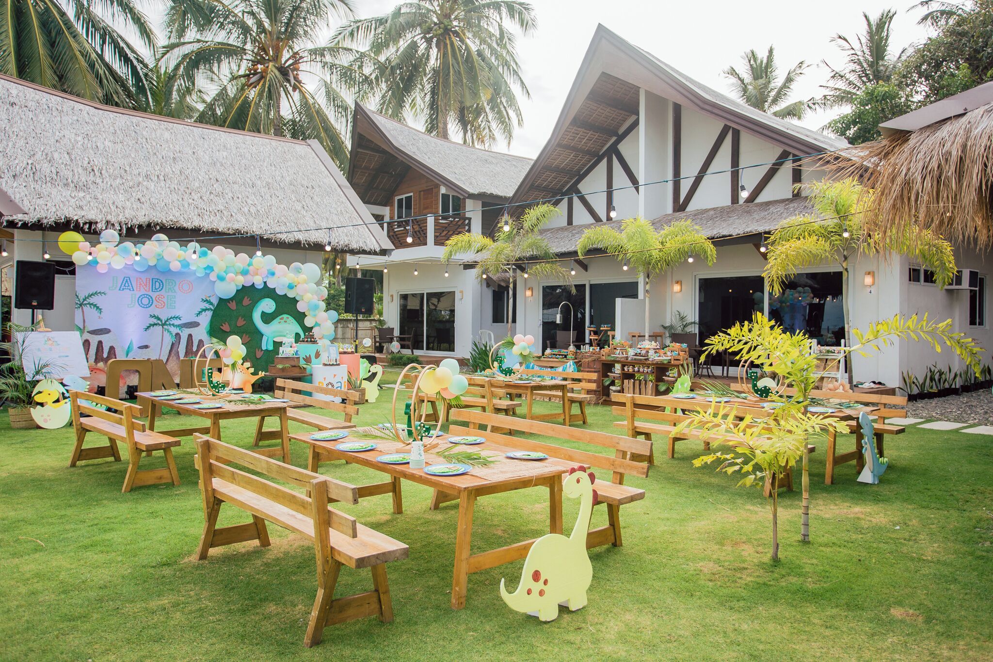 An outdoor dining area with wooden tables and benches, surrounded by palm trees and a building with a thatched roof.