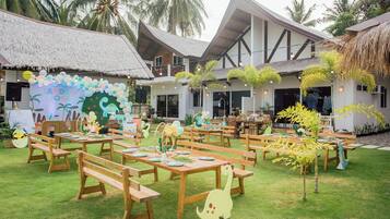 An outdoor dining area with wooden tables and benches, surrounded by palm trees and a building with a thatched roof.