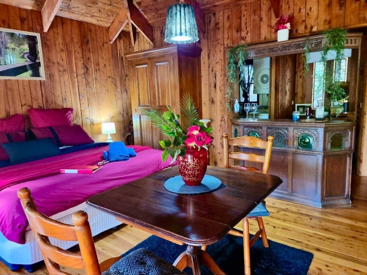 A wooden cabin interior with a bed, a dining table with a vase of flowers, and a kitchen area.