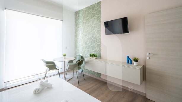 A modern hotel room with a large window, a flat-screen TV mounted on the wall, a small round table with two chairs, a white cabinet with a plant, and a wooden door. - Homiday - Residence Venere, Interior 6 (Alba Adriatica)