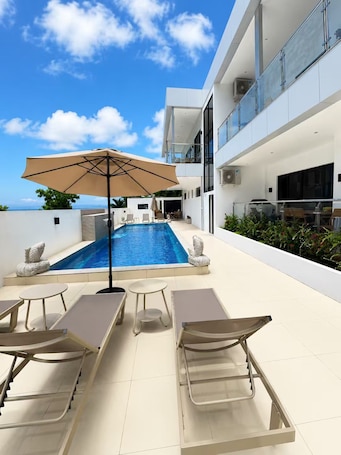 A modern outdoor pool area with lounge chairs, a large umbrella, and a clear view of the sky.
