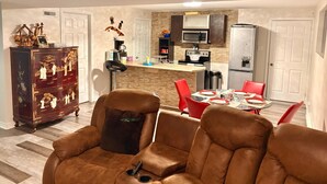 A modern living room with a brown leather sofa, a dining area with red chairs, and a kitchen with a microwave and refrigerator.