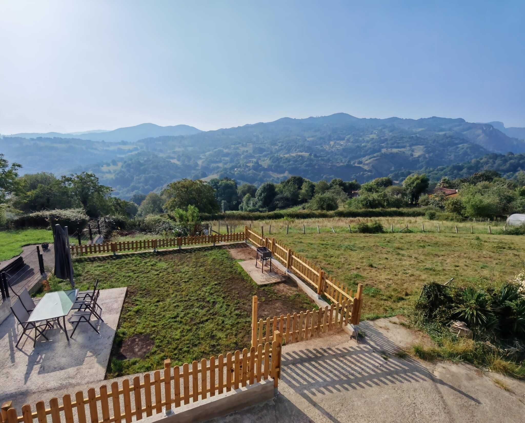 Un espace extérieur clos, aménagé avec une table et des chaises, entouré d’un paysage vallonné et de montagnes lointaines.