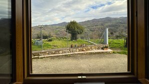 Vista desde una ventana que muestra un muro de piedra, un árbol y un paisaje montañoso.