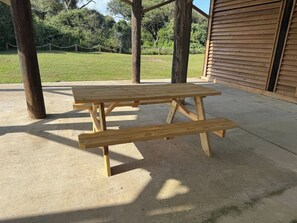 Una mesa de picnic de madera con bancos en un espacio exterior con piso de concreto y un edificio de madera al fondo.