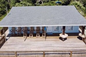 Un espacio de estar al aire libre cubierto, con sillas y una mesa de madera, una terraza de madera y un edificio con techo gris.