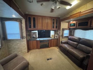 A compact RV interior with a kitchenette, a brown leather couch, and a ceiling fan.