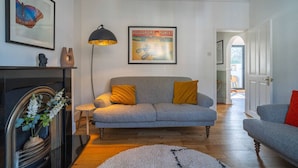 Een woonkamer met een grijze bank, een zwarte vloerlamp, een ingelijst poster en een open haard met decoratieve objecten.