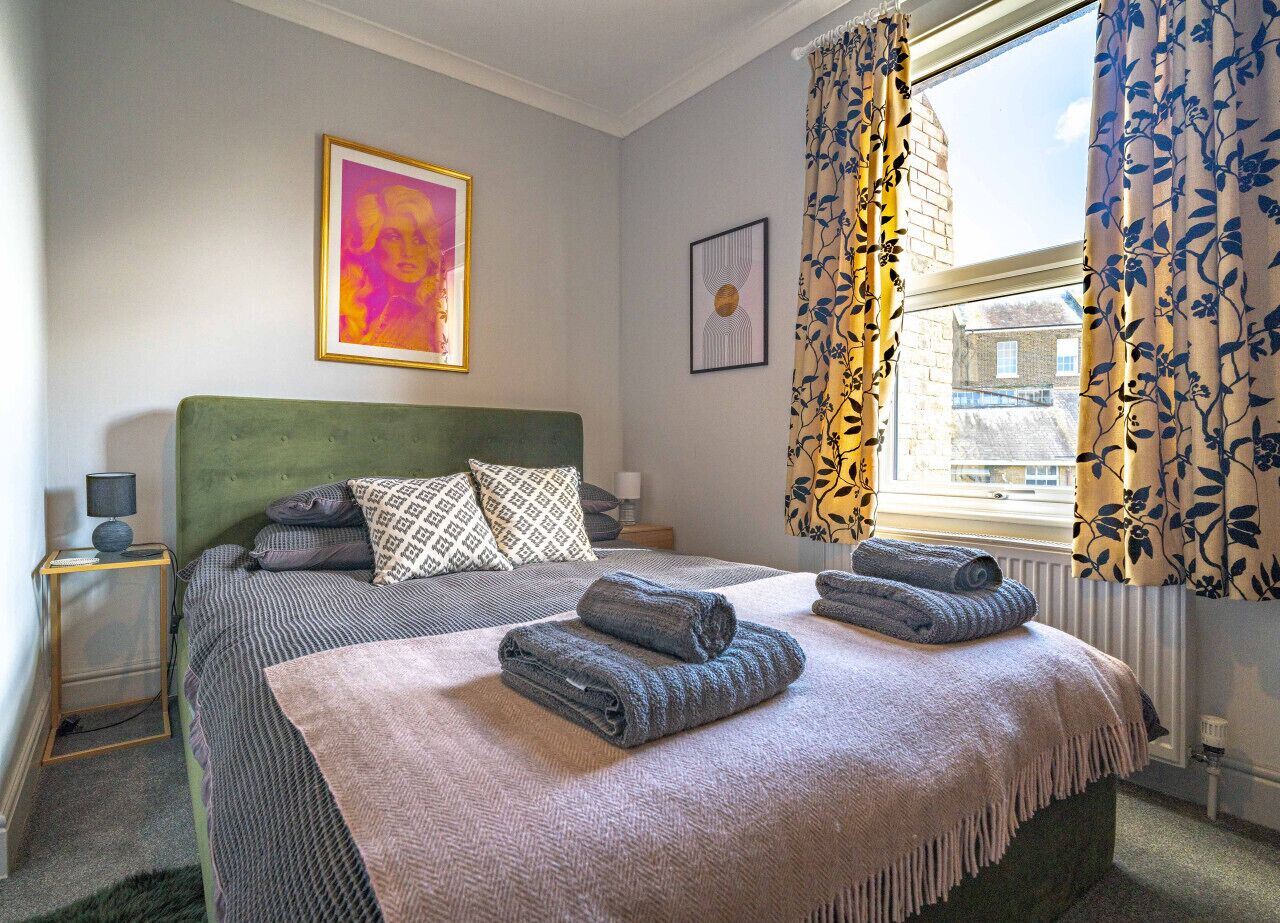 Un lit bien fait, avec une tête de lit verte, deux oreillers et des serviettes pliées. Un tableau encadré est accroché au mur, il y a une fenêtre avec des rideaux à motifs floraux et une petite table de chevet avec une lampe.