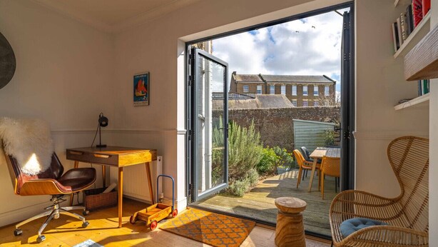 Een kamer met een grote schuifglazen deur die uitkomt op een terras met een tafel en stoelen, een rieten stoel, een houten bureau met stoel en een radiator.