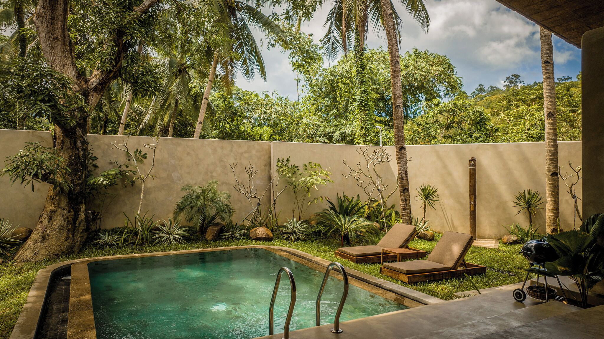 A pool area with lounge chairs, a barbecue, and tropical plants.