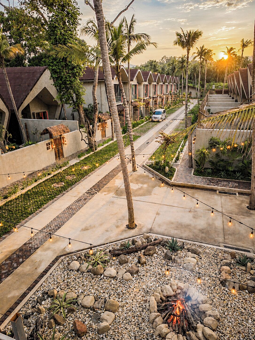A resort with a fire pit area, palm trees, and a row of houses with a sunset in the background.