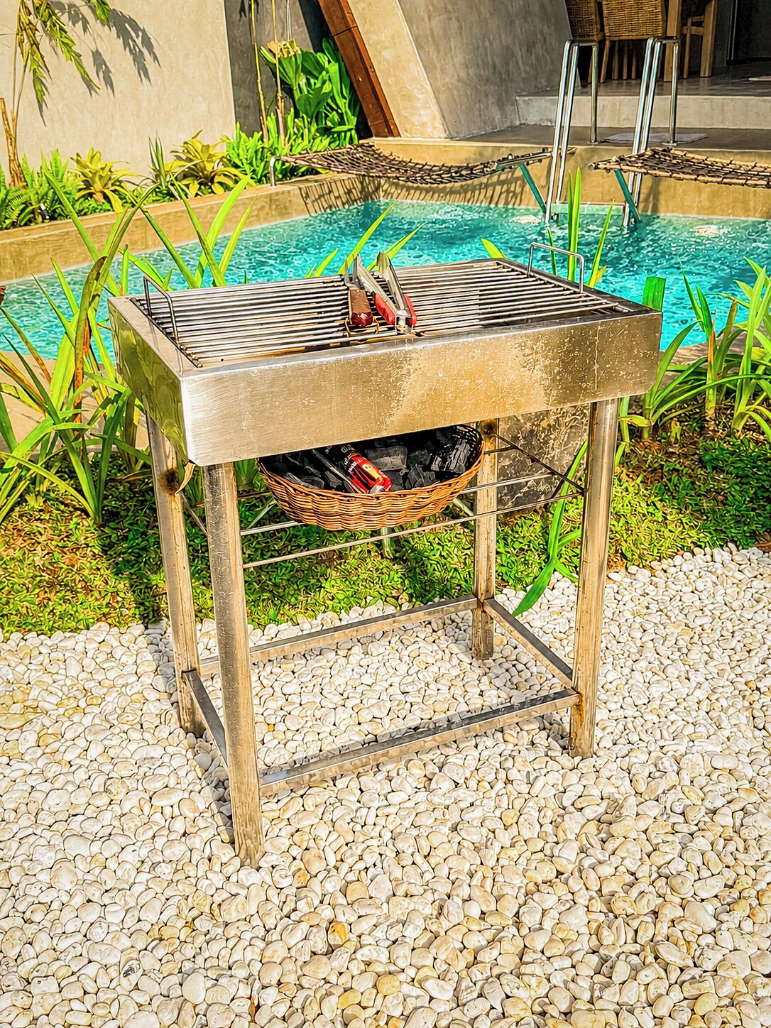 A portable barbecue grill with a basket underneath, situated on a pebbled surface near a swimming pool.