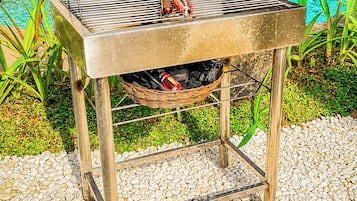 A portable barbecue grill with a basket underneath, situated on a pebbled surface near a swimming pool.