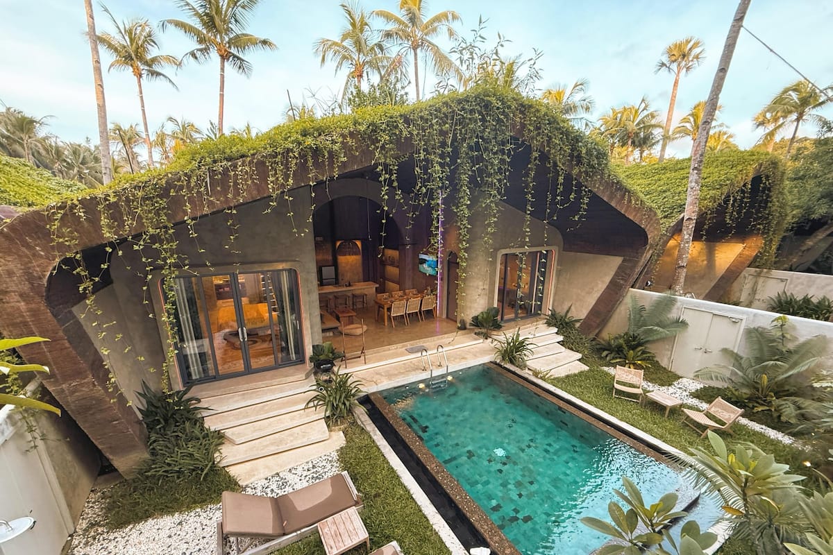 Un área de piscina moderna al aire libre con solárium, rodeada de palmeras y una zona de estar cubierta.
