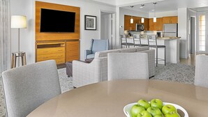 Ein modernes Hotelzimmer mit Essbereich, einem wandmontierten Flachbildfernseher, einer Küchenzeile mit Holzschränken und einer Sitzecke mit rundem Tisch und Stühlen.
