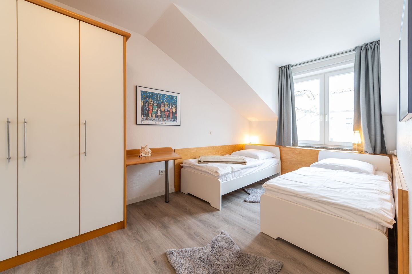 A bedroom with two beds, a desk, a wardrobe, and a window with curtains.