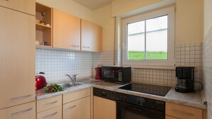 A kitchen with light wooden cabinets, a stainless steel sink, a red kettle, a black microwave, and a coffee maker.