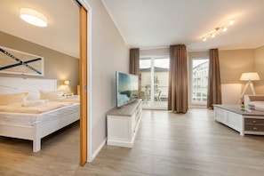 A modern hotel room with a large bed, a TV, a desk, and a balcony with curtains. - Residence Bel Vital - Apartment 12 Bella Vita - Apartment Bella Vita im (Binz)