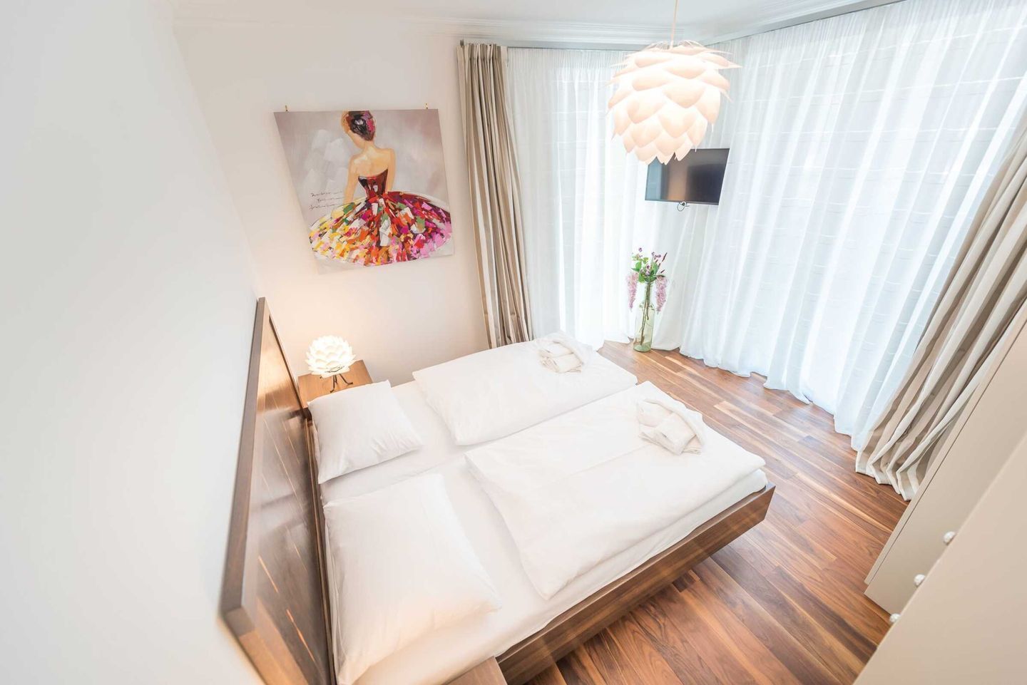 Ein Hotelzimmer mit einem großen Bett, Holzboden, einem Gemälde an der Wand, einem an der Wand befestigten Fernseher und einer hängenden Leuchte.