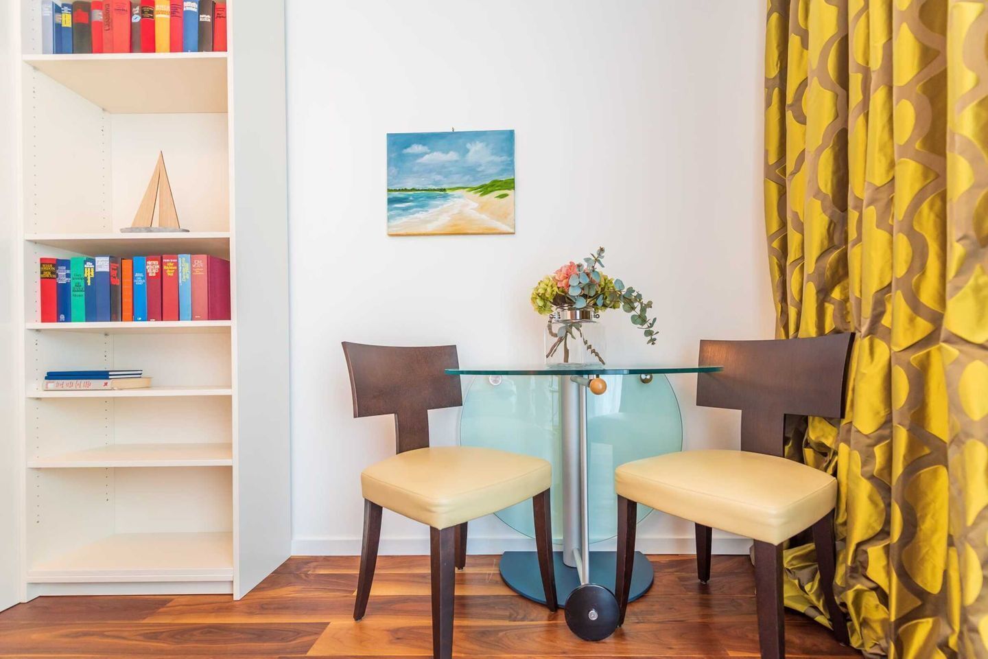 Une pièce avec une table en verre, deux chaises, une bibliothèque, un tableau et un rideau jaune.
