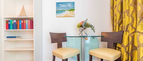 Une pièce avec une table en verre, deux chaises, une bibliothèque, un tableau et un rideau jaune.