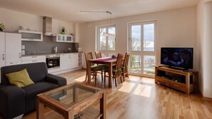Ein modernes Wohnzimmer mit Sofa, Couchtisch, Esstisch, Stühlen, Fernseher, Küchenzeile und großen Fenstern.