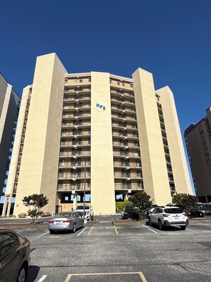 Un immeuble de grande hauteur, dont la façade est ornée d’un logo HPS bien visible, avec de nombreux balcons et un parking situé au premier plan.