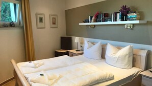 Ein Hotelzimmer mit Bett, Nachttisch, einer Ablage mit Büchern und Blumen sowie einem Fenster mit Vorhängen.