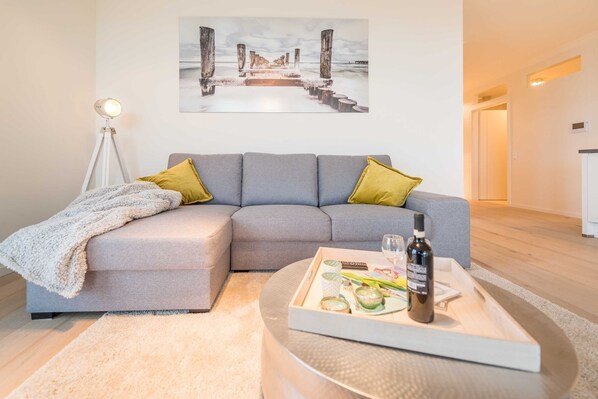 A modern living room with a grey sectional sofa, a coffee table with a bottle and glasses, a painting of a pier, and a floor lamp.