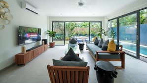 A modern living room with a TV, seating, and a view of a pool. - Twin Villas Drupadi 8Br Seminyak (Seminyak)