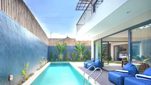 A modern outdoor pool area with a clear blue pool, a wooden fence, and a glass door leading to a room with a bed and a chair. - Twin Villas Drupadi 8Br Seminyak (Seminyak)
