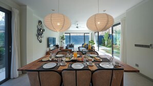 A dining area with a large table set for a meal, surrounded by wicker chairs, with a view of a pool and greenery outside. - Twin Villas Drupadi 8Br Seminyak (Seminyak)