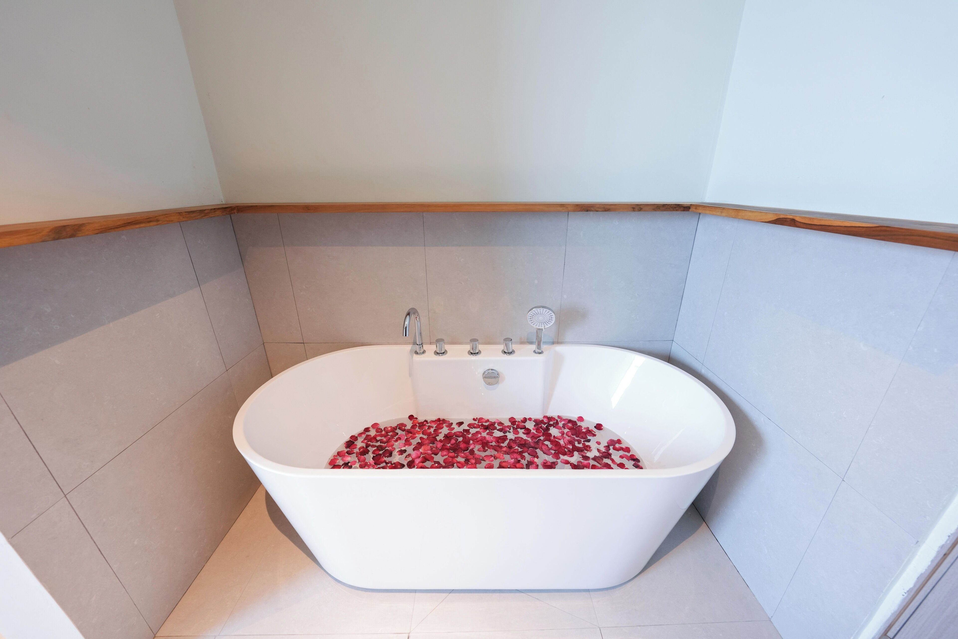 A modern bathroom with a freestanding bathtub filled with rose petals, a wooden floor, and a tiled wall.