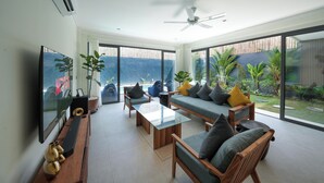 A modern living room with a flat-screen TV, wooden furniture, and large windows overlooking a garden. - Twin Villas Drupadi 8Br Seminyak (Seminyak)