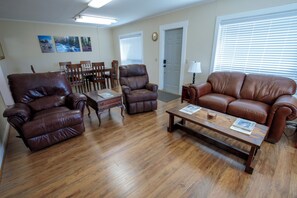 A living room with a brown leather sofa, a brown leather armchair, a wooden coffee table, and a dining area with wooden chairs and a table.
