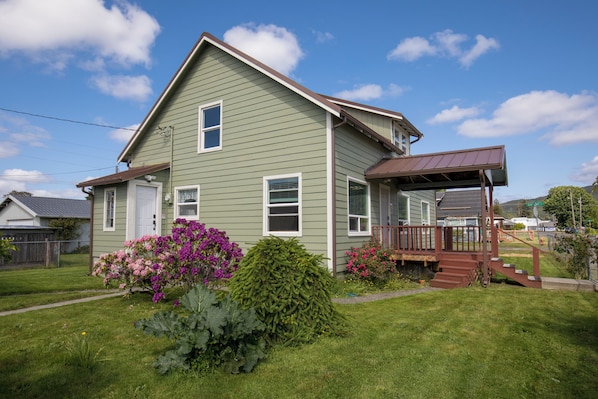 A two-story house with a front porch, a small garden with flowers, and a clear sky.
