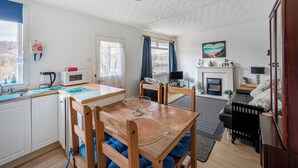 A cozy living room with a fireplace, a dining area with a wooden table and chairs, and a kitchen with white cabinets and a window. - 2 Bed Chalet - Sleeps 4 - River and mountain views (Strathpeffer)