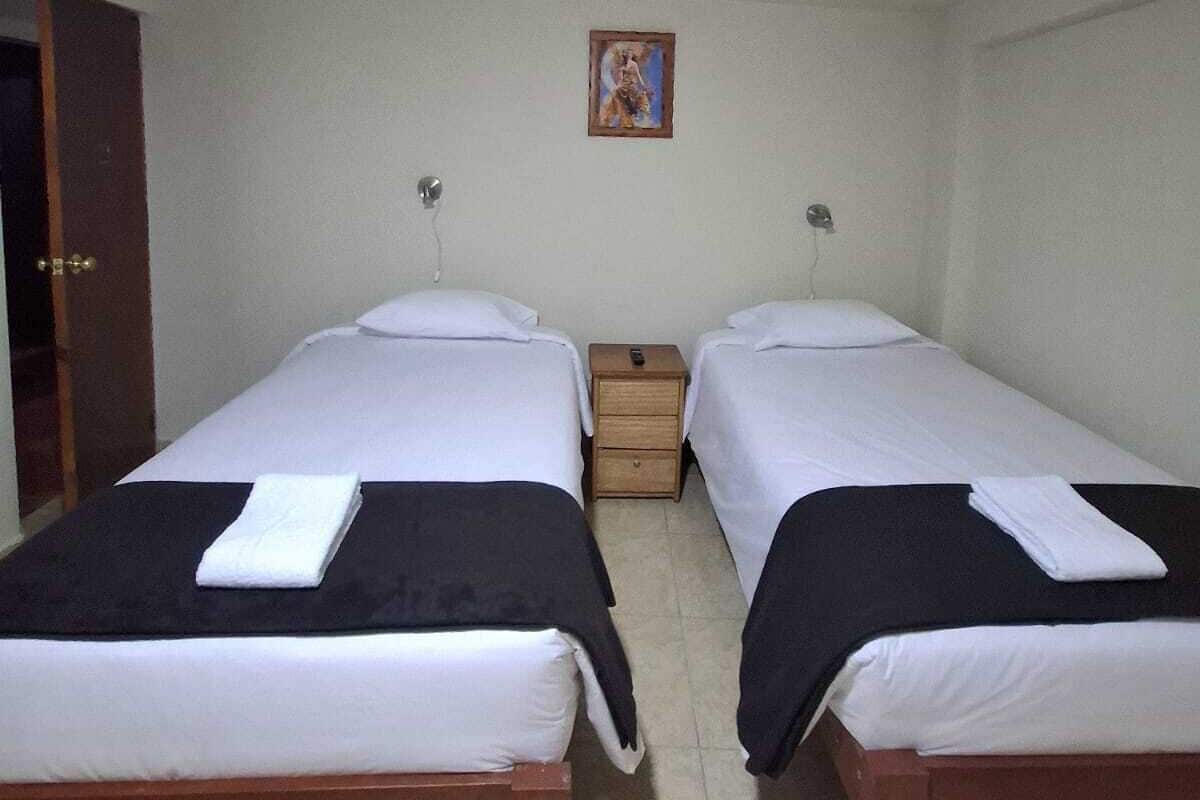 Une petite chambre propre, avec deux lits simples, une table de chevet en bois, une petite table et une porte.