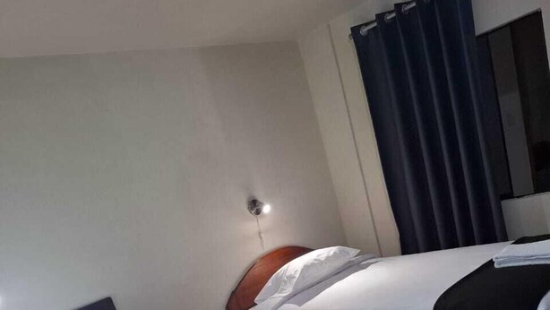 Habitación de hotel con dos camas, cada una con ropa de cama blanca y cabeceros de madera.  Entre las camas hay una mesita con una toalla doblada. La habitación tiene una cortina oscura en un lado y una lámpara fijada a la pared.