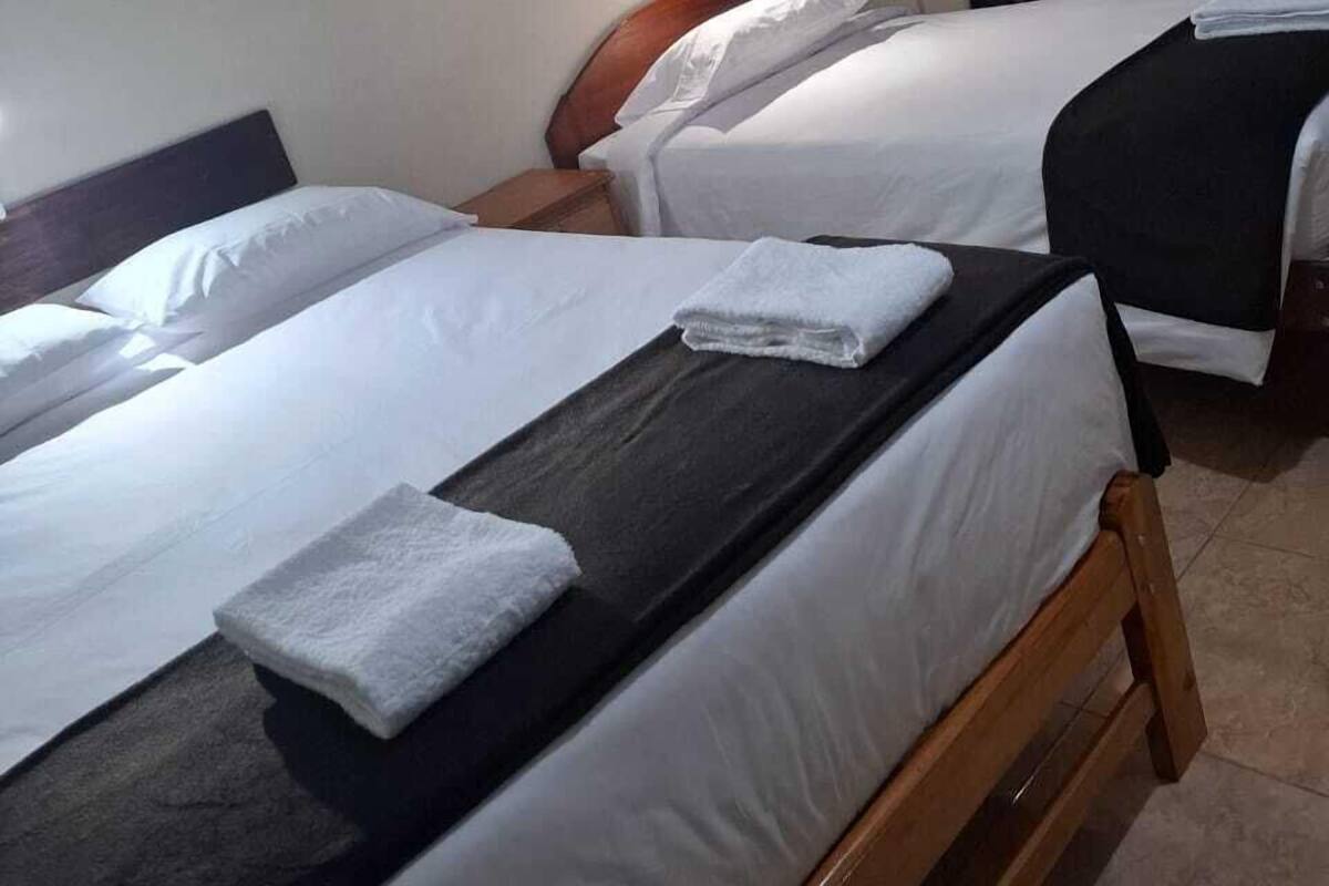 Une chambre d’hôtel avec deux lits, chacun doté de draps blancs et d’une tête de lit en bois. Une petite table se trouve entre les lits, sur laquelle est posée une serviette pliée. La chambre est équipée d’un rideau foncé d’un côté et d’un luminaire fixé au mur.
