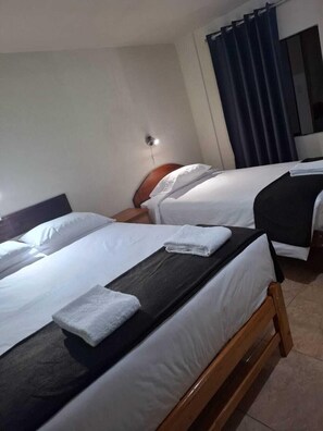 A hotel room with two beds, each with white linens and a wooden headboard. There is a small table between the beds with a folded towel on it. The room has a dark curtain on one side and a wall-mounted light fixture.