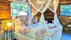 A wooden cabin room with a bed under a mosquito net, a nightstand with a lamp, and a stone wall. - Forget Me Not Cottages-Rose Cottage (Warburton)